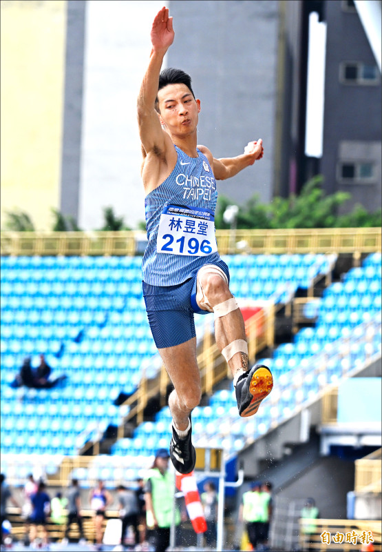 台灣跳遠選手林昱堂在2025台灣田徑公開賽中起跳瞬間，身穿藍色戰袍、空中動作有力，背景為藍色觀眾席與田徑場。