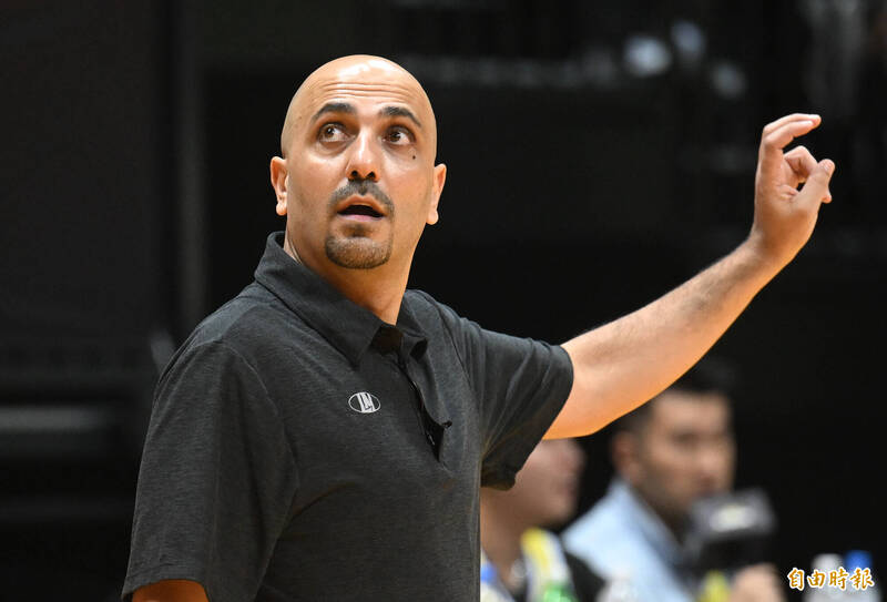 一名籃球教練身穿深色 POLO 衫在場邊指揮比賽，表情專注，背景為模糊的觀眾席。