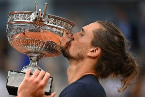 2026年法網男單冠軍茲維列夫（Alexander Zverev）留著招牌金色長髮並紮成髮髻，身穿深藍色網球比賽球衣。他閉上雙眼，神情無比深情且充滿敬意地用雙手捧起神聖的法網火槍手冠軍獎盃，並在獎盃光滑的金屬表面上落下一吻，古銅色紅土球場的倒影清晰地映照在盃身之上。構圖神韻請參考 "php7ZTFF3.jpg"。