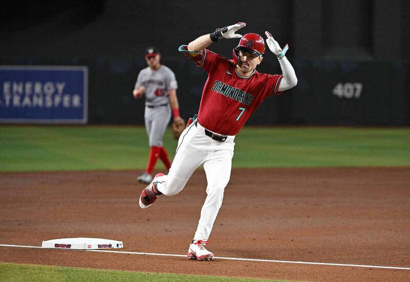 亞利桑那響尾蛇隊球星卡洛爾（Corbin Carroll）身穿紅色「DIAMONDBACKS 7」字樣的經典主場球衣、白色球褲、頭戴紅色亮面打擊頭盔，在繞壘時面向隊友休息區高高躍起，雙手舉至頭頂兩側做出招牌的慶祝動作。畫面後方可見身穿灰色客場球衣的國民隊野手黯然走過。構圖神韻可以參考 "phpaQSY2r.jpg"。