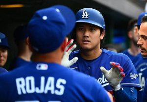 道奇隊日本超級巨星大谷翔平在檔案 "phpWxuiM1.jpg" 中，身穿道奇隊經典的皇家藍客場比賽球衣，頭戴印有白色「LA」隊徽的亮藍色打擊頭盔。他在敲出本季第 13 轟後回到休息區（Dugout），正揚起嘴角帶著自信且放鬆的微笑，雙手戴著白底配紅藍線條的防護打擊手套高高舉起，準備與前方身穿背號「ROJAS 11」球衣的隊友羅哈斯（Miguel Rojas）以及右側神情興奮的隊友們一一擊掌慶祝，生動捕捉了全壘打英雄凱旋回歸休息室的榮耀瞬間。