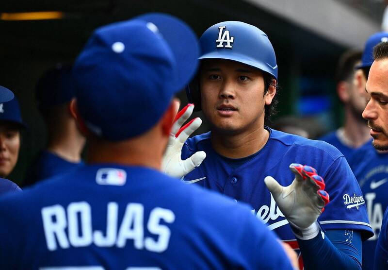 道奇隊日本超級巨星大谷翔平在檔案 "phpWxuiM1.jpg" 中，身穿道奇隊經典的皇家藍客場比賽球衣，頭戴印有白色「LA」隊徽的亮藍色打擊頭盔。他在敲出本季第 13 轟後回到休息區（Dugout），正揚起嘴角帶著自信且放鬆的微笑，雙手戴著白底配紅藍線條的防護打擊手套高高舉起，準備與前方身穿背號「ROJAS 11」球衣的隊友羅哈斯（Miguel Rojas）以及右側神情興奮的隊友們一一擊掌慶祝，生動捕捉了全壘打英雄凱旋回歸休息室的榮耀瞬間。