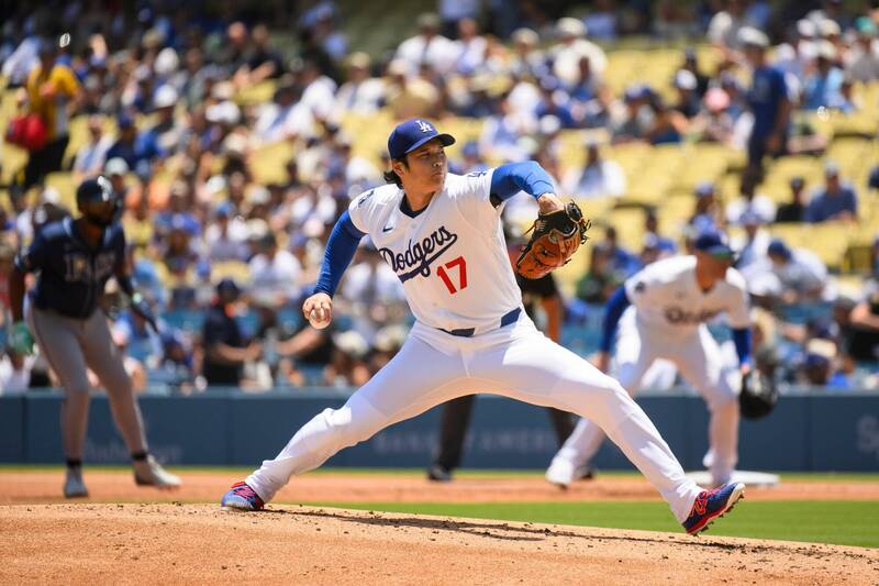 洛杉磯道奇隊日本球星大谷翔平身穿白色 17 號主場球衣、戴著藍色球帽，在投手丘上展現標誌性的右投出手投球動態，雙腿大跨步拉開、延伸極具張力，背景為看台上密密麻麻的道奇球迷。