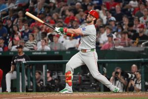 費城人隊打者哈波（Bryce Harper）身穿灰色客場球衣、戴著紅色打擊頭盔與綠色防護具，在球場上維持打擊完隨球棒揮過身體的延伸姿勢，一邊抬頭目送全壘打飛出場外。