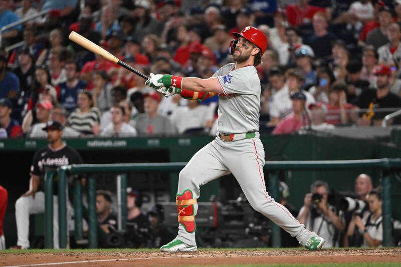 費城人隊打者哈波（Bryce Harper）身穿灰色客場球衣、戴著紅色打擊頭盔與綠色防護具，在球場上維持打擊完隨球棒揮過身體的延伸姿勢，一邊抬頭目送全壘打飛出場外。
