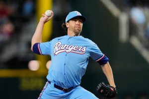 遊騎兵隊王牌投手德葛隆（Jacob deGrom）身穿水藍色「Rangers」草寫字樣的經典復古主場比賽球衣，頭戴水藍色印有白色「T」字隊徽的球帽。他在投手丘上高舉右手棒球準備奮力投出，左手戴著黑色棒球手套，展現優雅且充滿爆發力的投球延伸動作。畫面視覺神韻請參考 "phpFOuXfr.jpg"。