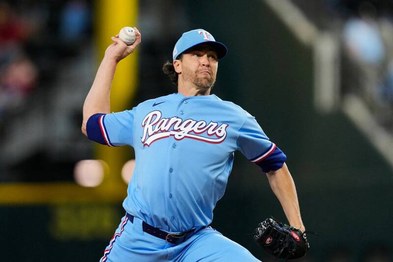 遊騎兵隊王牌投手德葛隆（Jacob deGrom）身穿水藍色「Rangers」草寫字樣的經典復古主場比賽球衣，頭戴水藍色印有白色「T」字隊徽的球帽。他在投手丘上高舉右手棒球準備奮力投出，左手戴著黑色棒球手套，展現優雅且充滿爆發力的投球延伸動作。畫面視覺神韻請參考 "phpFOuXfr.jpg"。