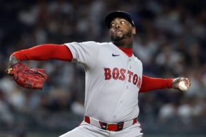 紅襪隊古巴火球男查普曼（Aroldis Chapman）在照片中，身穿紅襪隊灰色的客場比賽球衣，胸前繡有紅色的「BOSTON」字樣，內搭紅色長袖緊身衣，戴著紅襪隊深藍色的經典球帽。他正身處球場投手丘上，展現其招牌且孔武有力的左投出手投球分解動作，他的右手戴著亮紅色的棒球手套向後大幅度擺盪延伸，左手則握著棒球全神貫注地準備向前全力的鞭甩投出，臉部神情因用力而顯得有些扭曲且極具專注度，背景是模糊的夜間球場外野看台燈火。