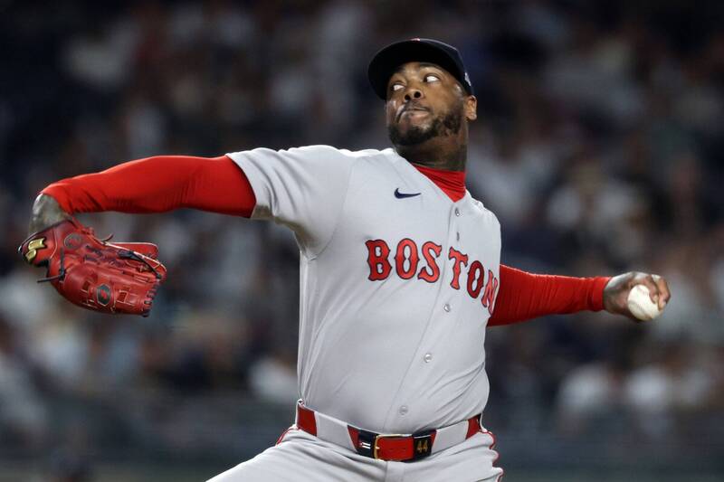 紅襪隊古巴火球男查普曼（Aroldis Chapman）在照片中，身穿紅襪隊灰色的客場比賽球衣，胸前繡有紅色的「BOSTON」字樣，內搭紅色長袖緊身衣，戴著紅襪隊深藍色的經典球帽。他正身處球場投手丘上，展現其招牌且孔武有力的左投出手投球分解動作，他的右手戴著亮紅色的棒球手套向後大幅度擺盪延伸，左手則握著棒球全神貫注地準備向前全力的鞭甩投出，臉部神情因用力而顯得有些扭曲且極具專注度，背景是模糊的夜間球場外野看台燈火。