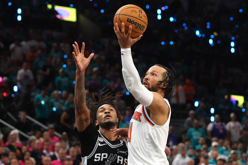 紐約尼克隊當家球星布朗森（Jalen Brunson）身穿白底橙藍邊的11號經典球衣、右臂套著白色防護袖套，在籃下躍起，雙手高舉威爾森（Wilson）品牌籃球準備在馬刺隊防守球員的封阻下完成拋投。
