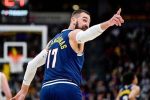 金塊隊中鋒瓦倫丘納斯(Jonas Valanciunas)身穿印有黃色「VALANCIUNAS」及 17 號字樣的深藍色金塊隊客場球衣。他留著標誌性的落腮鬍,雙臂套著白色長袖護臂,正轉過頭並伸出右手食指指向遠方球場,向隊友或裁判示意。