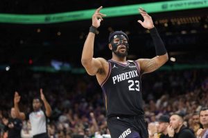 太陽隊後衛古德溫(Jordan Goodwin)在球場檔案 "phpAYDzJF.jpg" 中,身穿太陽隊黑色 23 號主題球衣,面部戴著黑色的防護面具。他雙臂高舉、雙手張開、神情專注地在比賽球場上慶祝或呼喊隊友,手腕與左手臂皆戴著黑色護腕與袖套。