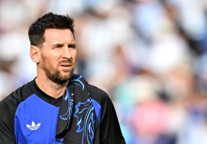 阿根廷球王梅西(Lionel Messi)蓄著整齊的落腮鬍,身穿黑藍雙色拼接、胸前印有經典三葉草愛迪達商標的國家隊運動訓練服,肩上披著一條帶有藍黑色圖騰的毛巾。他站在球場上,目光直視前方,神情顯得有些嚴肅與專注,背景為有些模糊的球場觀眾席。