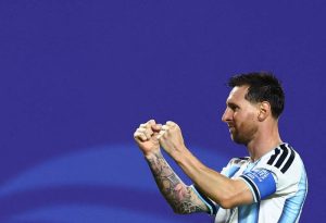 阿根廷球王梅西(Lionel Messi)在照片檔案 "php5RM7ZH.jpg" 中,身穿經典藍白條紋國家隊球衣,右手大片刺青清晰可見,在藍色背景前側身面向右方、雙手握拳興奮慶祝。