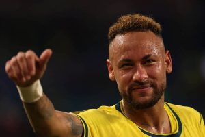 巴西球星內馬爾（Neymar）留著金色短捲髮、身上帶著汗水，面帶微笑並舉起右手大拇指比讚，身穿黃色綠領的巴西國家隊球衣。
