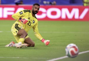 沙烏地阿拉伯門將奧維斯(Mohammed Al Owais)身穿 21 號亮黃色球衣與紅白守門手套,在世界盃綠色草皮球場上完成飛撲防守後,雙膝跪地並全神貫注地盯著滾動中的足球。